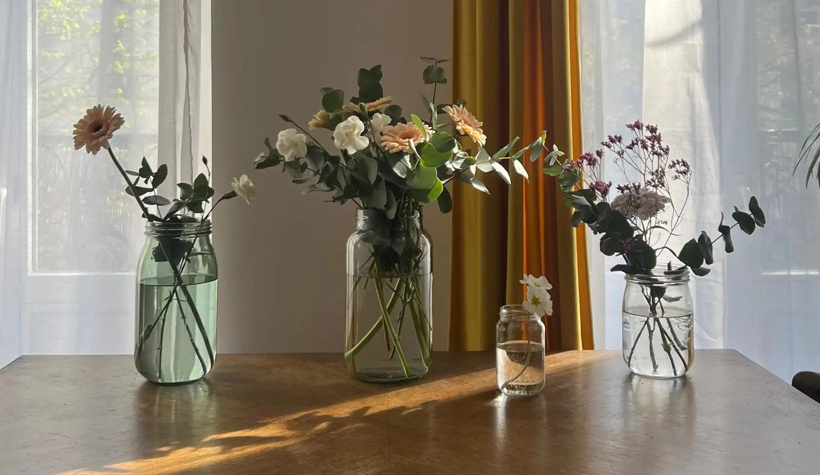 Trois bouquets de fleurs dans des bocaux sur une table en bois, devant des rideaux de lumière
