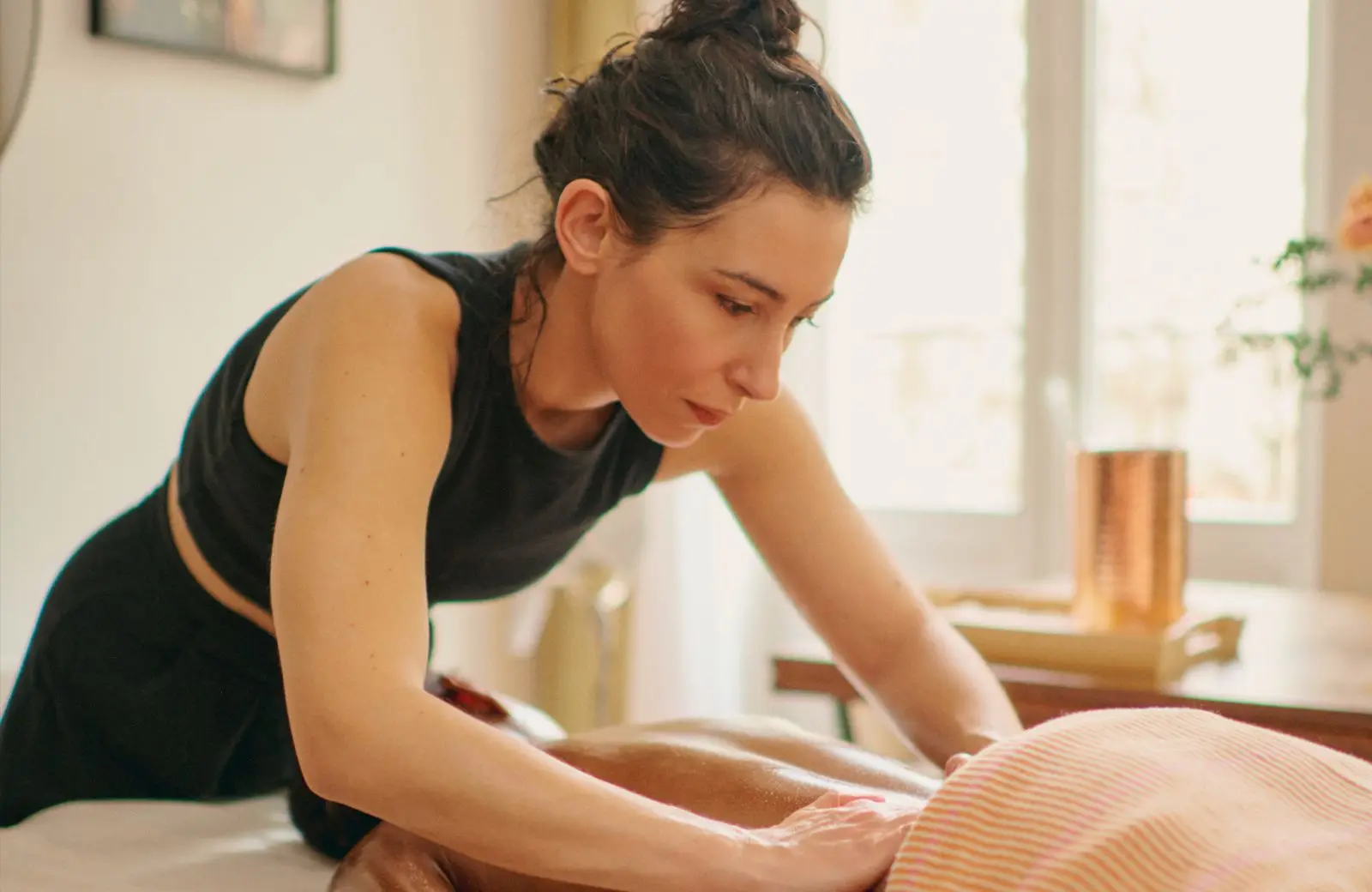 Clémence penchée sur le dos d’une cliente pendant un massage, concentrée, dans la lumière d’une fenêtre