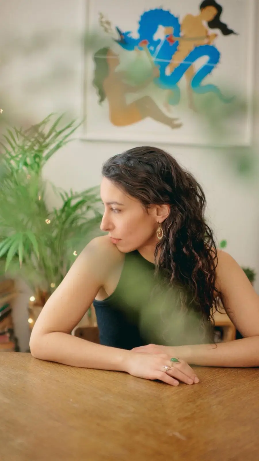 Clémence, accoudée à une table en bois, le regard tourné vers la gauche, entre des plantes et une grande illustration colorée